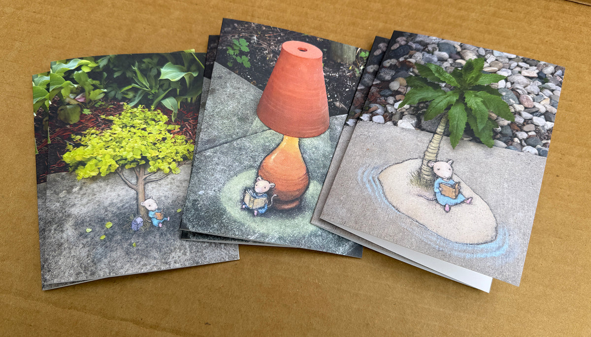 Six illustrated greeting cards fanned out on a tan surface. The cards show whimsical garden scenes: one with a bright yellow-green tree, one with a terracotta lamp, and one with a small palm tree on a sandy island.