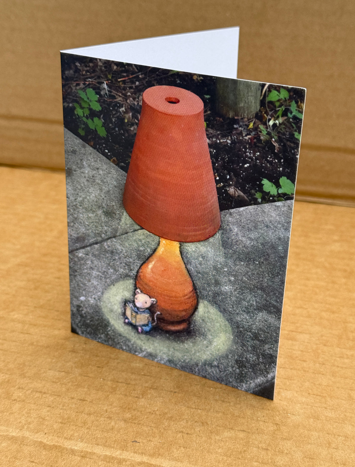 A folded greeting card standing upright on a cardboard background. The card features a whimsical illustration of a chalk drawing of a lamp with an overturned flower pot as its shade, with a small mouse sitting at the base reading a book.