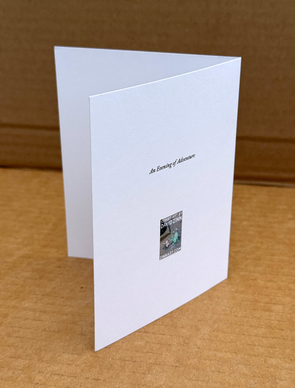 A white folded greeting card standing open on a brown cardboard surface, with printed text reading "An Evening of Adventure"