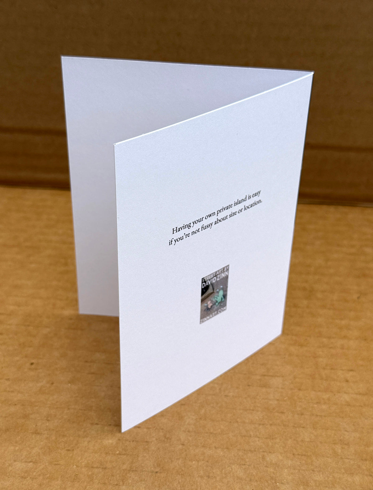A white folded greeting card standing open on a brown cardboard surface, with printed black text reading "Having your own private island is easy if you're not fussy about size or location"