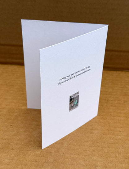 A white folded greeting card standing open on a brown cardboard surface, with printed black text reading "Having your own private island is easy if you're not fussy about size or location"