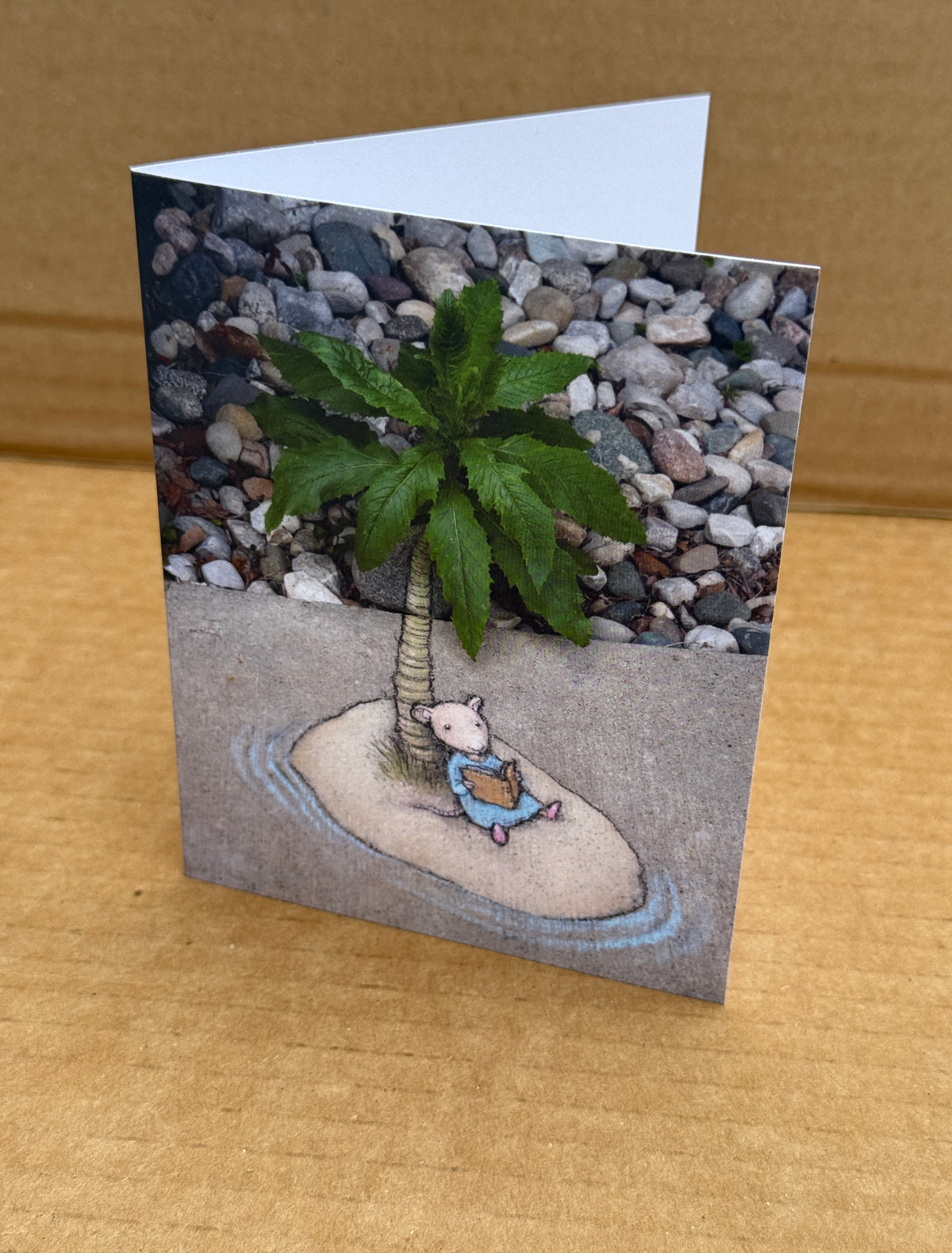 A folded greeting card standing upright on a cardboard background, printed with a whimsical photo of a small mouse sitting under a palm tree (the top of which is a garden weed) and reading a book on a desert island.