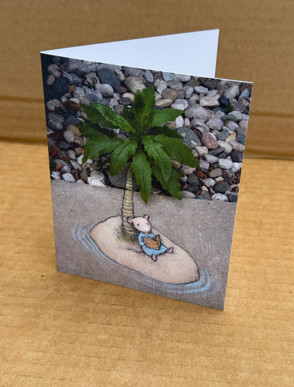 A folded greeting card standing upright on a cardboard background, printed with a whimsical photo of a small mouse sitting under a palm tree (the top of which is a garden weed) and reading a book on a desert island.